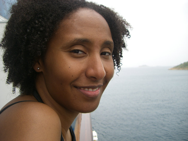 Close-up headshot of a smiling woman on a boat facing the camera with open water and small slivers of land in the background.