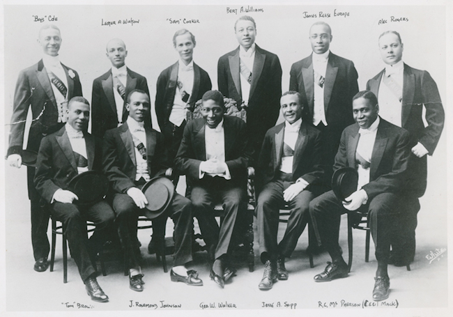 A group of gentleman dressed in suits, some holding hats, are pictured in two rows. Known as "The Frogs" the back row stands, the front row sits, and writing above or below each person identifies who they are.. 