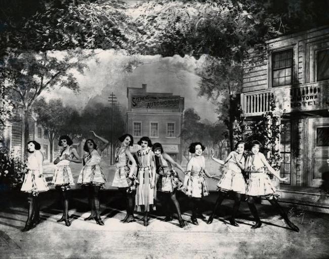 Production still from "Shuffle Along." Nine women in costume pose on stage in front of the set.