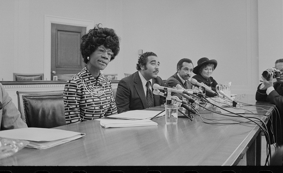 照片显示众议员雪莉·奇泽姆 (Shirley Chisholm)、众议员帕伦·米切尔 (Parren Mitchell)、众议员查尔斯·兰格尔 (Charles Rangel) 和众议员贝拉·阿布祖格 (Bella Abzug) 坐在带麦克风的桌子旁