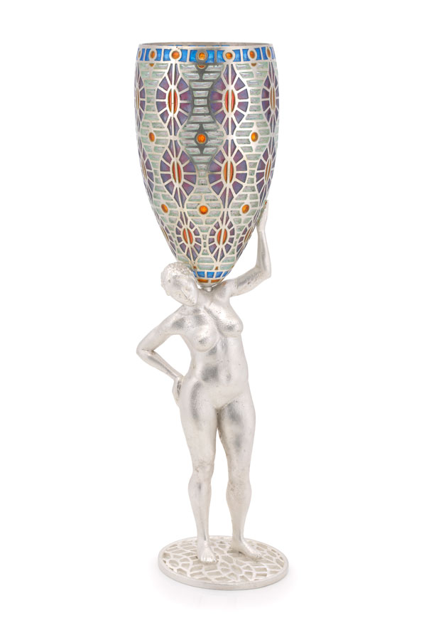 A tall wine glass with aa colored painted pattern on the glass. The stem of the glass is a silver statuette of a naked woman