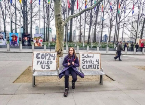 Alexandria Villaseñor sits in front of the UN in New York City. She sits on a silver bench in between to handmade signs. One says “School Strike 4 climate” and the other reads “COP24 Failed Us.” 