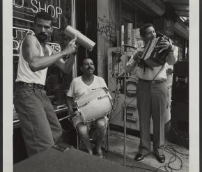 Fotografía en blanco y negro de tres hombres tocando instrumentos musicales fuera de una tienda. Un hombre está sentado con un tambor en el regazo, mientras que los otros dos están de pie: uno rasguea una güira y el otro sonríe mientras toca su instrumento.