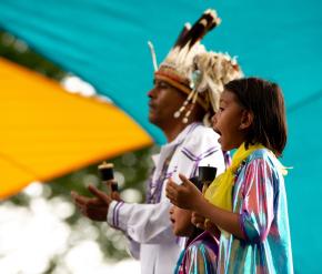 Un homme et une jeune fille en costume traditionnel chantent devant un fond turquoise.