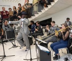 Dandy Wellington, a Black man in a gray striped suit dances in front of his seven piece jazz band in the Museum's rotunda. Behind them, guests in 1920s dress enjoy the performance. 