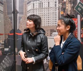Two people in front of a photograph of New York City, looking at exhibition objects
