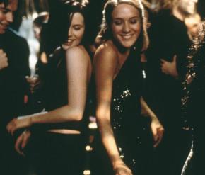 Kate Beckinsale and Chloë Sevigny smile and dance in black dresses in a crowded room. 