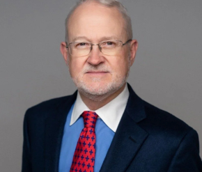 A man in a blue suit and red tie smiling in front of a grey background 