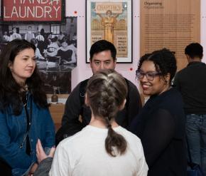 A crowd stands in a gallery with photographs, posters, and objects on pink walls