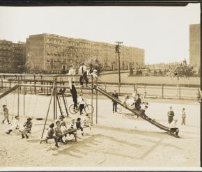 1921年に撮影された白黒写真。ブランコ、滑り台、シーソーのある遊び場で遊ぶ複数の子供たちが写っている。遊び場の後ろには通りとアパートの建物が見える。