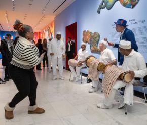 Three musicians wearing all white sitting on chairs playing latin drums. Women dancing in front of musicians.Artist Manny Vega in blue suit with shaker instrument.
