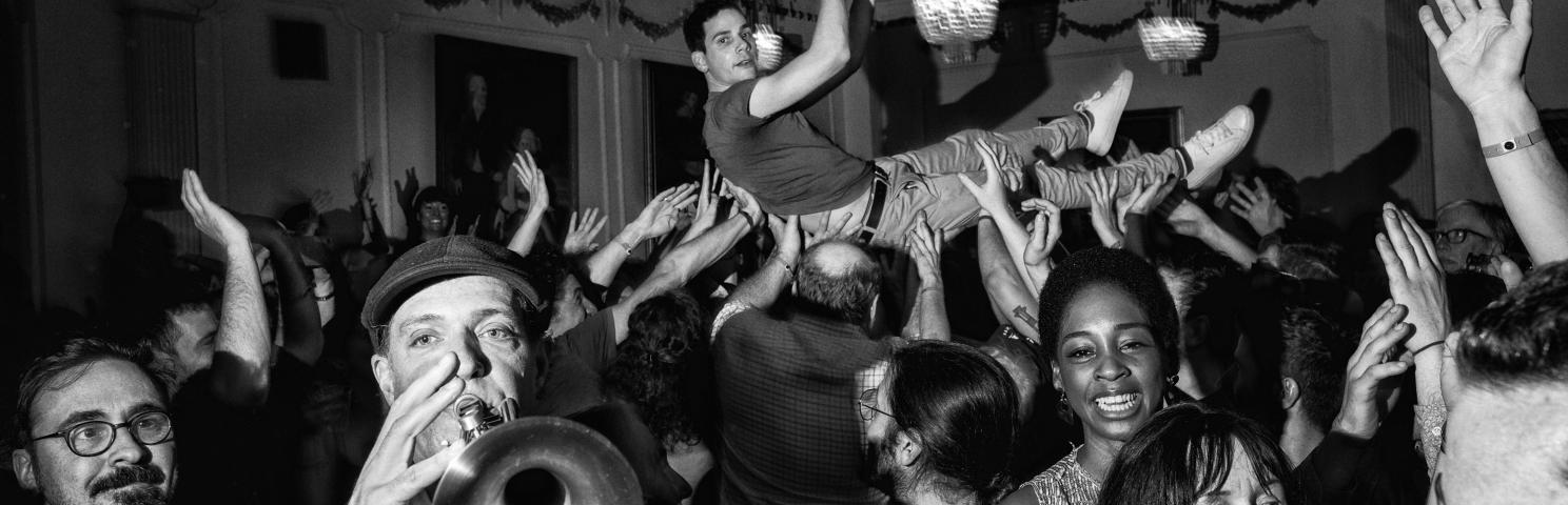 A crowd of people, including two people playing instruments in the foreground and a man crowdsurfing in the background