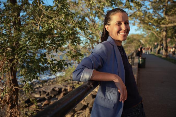 Tiffany Cabán sonríe y se inclina frente al puente de un parque. Lleva una chaqueta gris azulada y una blusa negra. Su cabello está recogido en una cola de caballo.