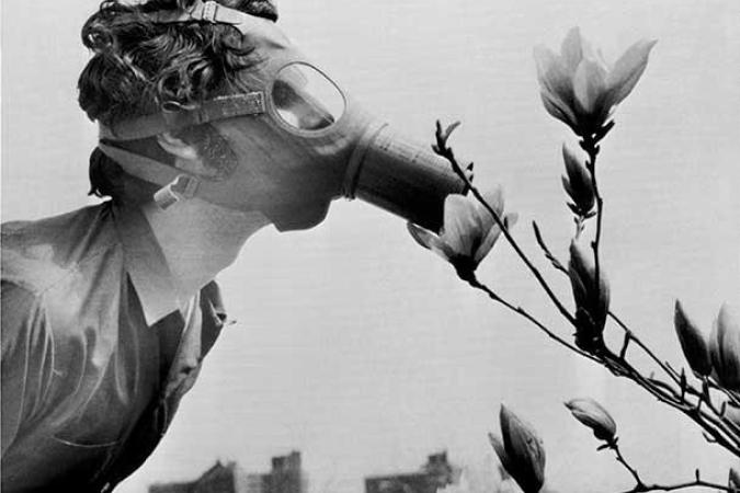 Fotografia em preto e branco de um homem usando uma máscara de gás inclinando-se para cheirar flores. Ao fundo, o horizonte de uma cidade.