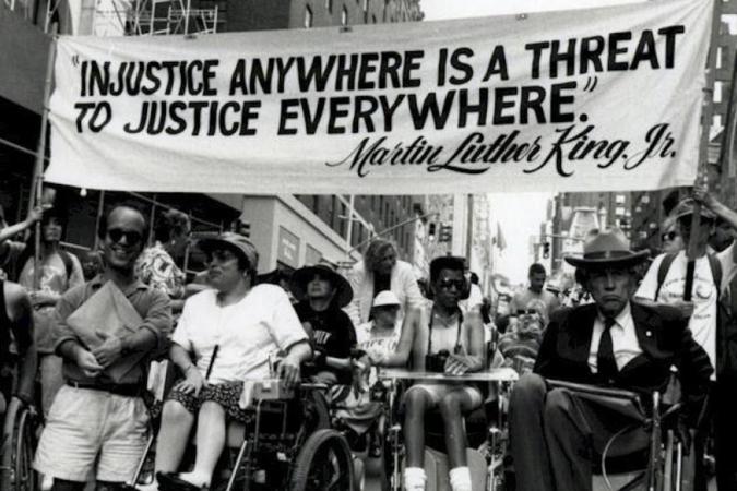 Une foule de personnes handicapées et de personnes en fauteuil roulant se rassemblent sous une banderole sur laquelle on peut lire "Injustice Anywhere est une menace pour la justice partout" Martin Luther King Jr.