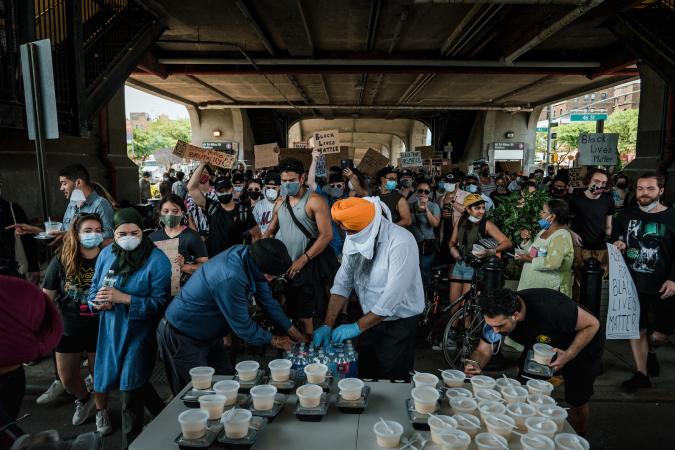 A crowd of people stand around a table with food containers. The man in the center is opening a case of water bottles to distribute.