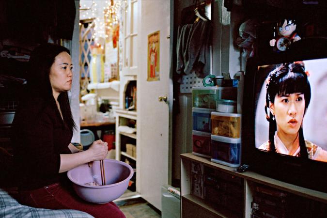 Una mujer china revuelve comida en un tazón mientras ve una telenovela china. Ella está sentada en una cama en un departamento.