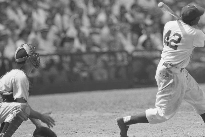 Jackie Robinson stands ready to bat in a crowded stadium as the pitcher and umpire look on from behind Robinson