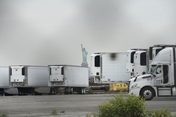 La Estatua de la Libertad representada detrás de camiones frigoríficos que sirven como depósito de cadáveres temporal para las víctimas de COVID