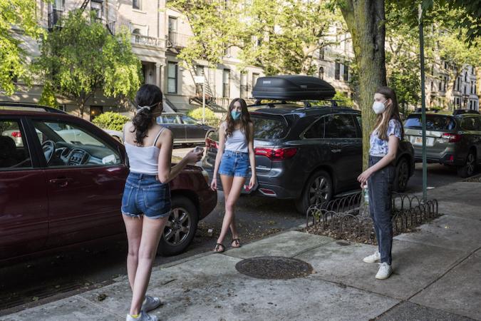 Three girls wearing masks stand apart but facing each other in a triangular fashion on the sidewalk in front of parked cars on a city side street
