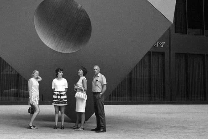 Four people stand on the sidewalk in front of a large statue of a cube with a hole going through the center of it