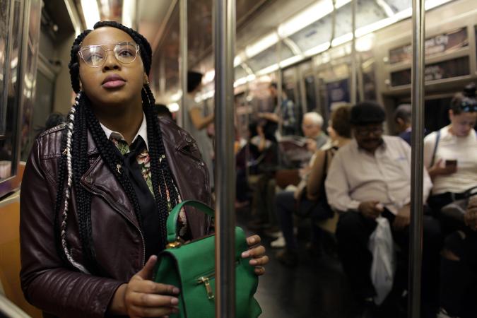 Imagem em close de mulheres de jaqueta roxa segurando uma sacola verde, sentadas em um trem do metrô de Nova York com outros passageiros.