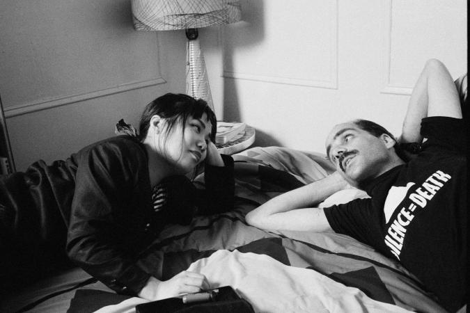 A female caregiver and male AIDS patient wearing an ACT UP t-shirt lie on a bed together looking into each other’s eyes 