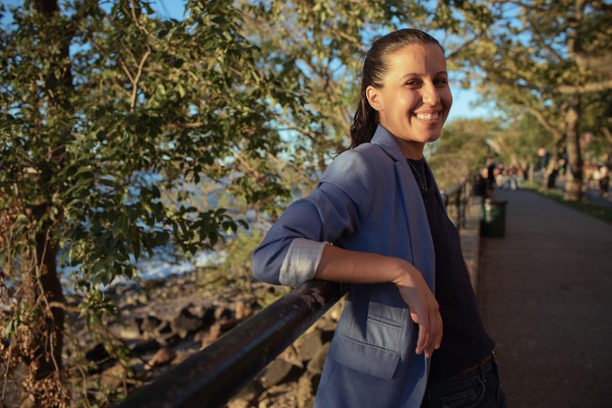Tiffany Cabán is smiling and leaning in front of a park bridge. She is wearing a bluish gray blazer and a black blouse. Her hair is tied back in a ponytail.