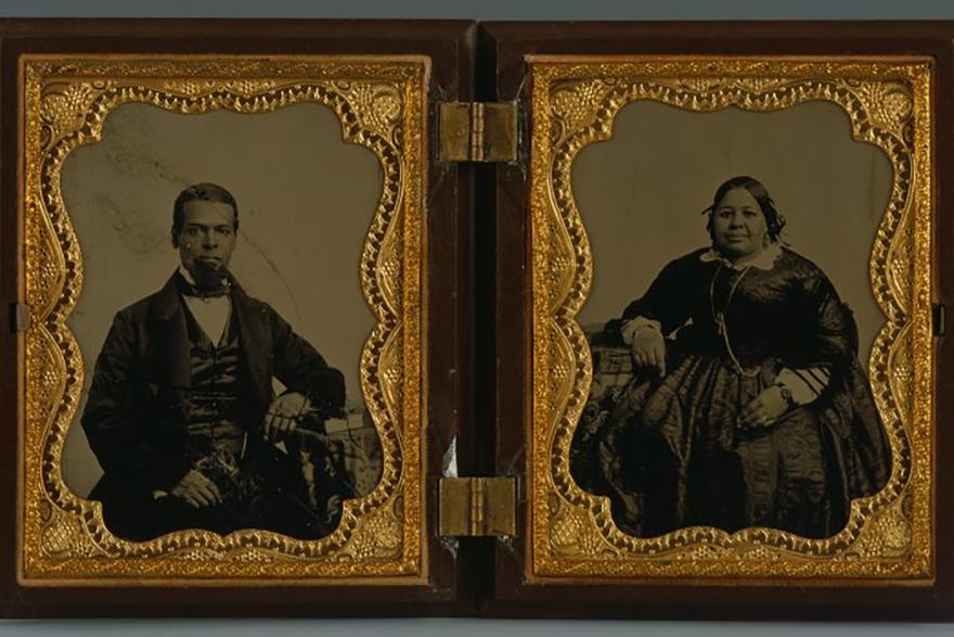 Two black and white photographs in a hinged wood and gold case. The man, left, and woman, right, are black and wear a fashionable dark suit and dress.
