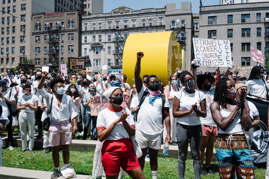 Uma multidão de manifestantes mestiços, mas predominantemente negros, se solidariza na praça do lado de fora do Museu do Brooklyn. Alguns seguram cartazes, outros levantam os punhos. Todos estão vestidos de branco.