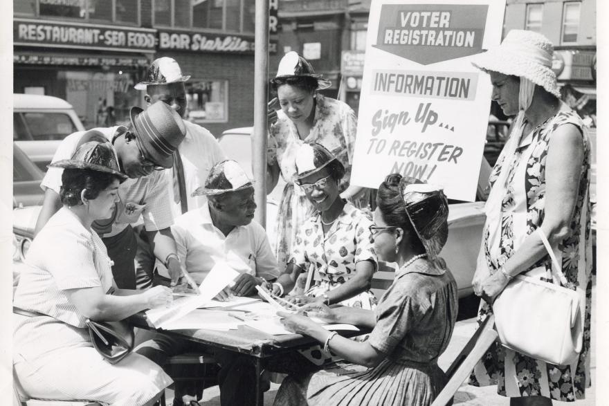 A black and white photograph of a group of Black men and women gathered around a table on the street with a sign that says Voter Registration behind them.