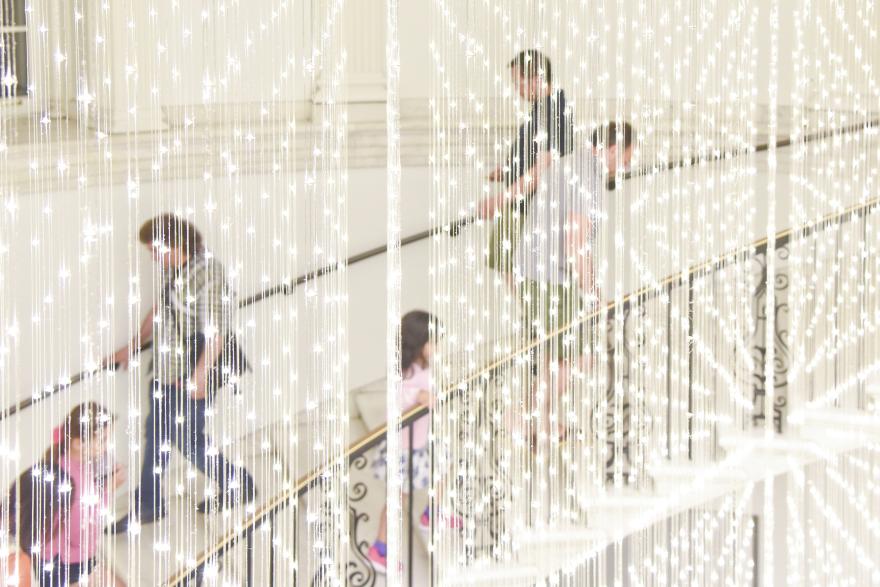 A crowd ascends a spiral staircase behind a light installation