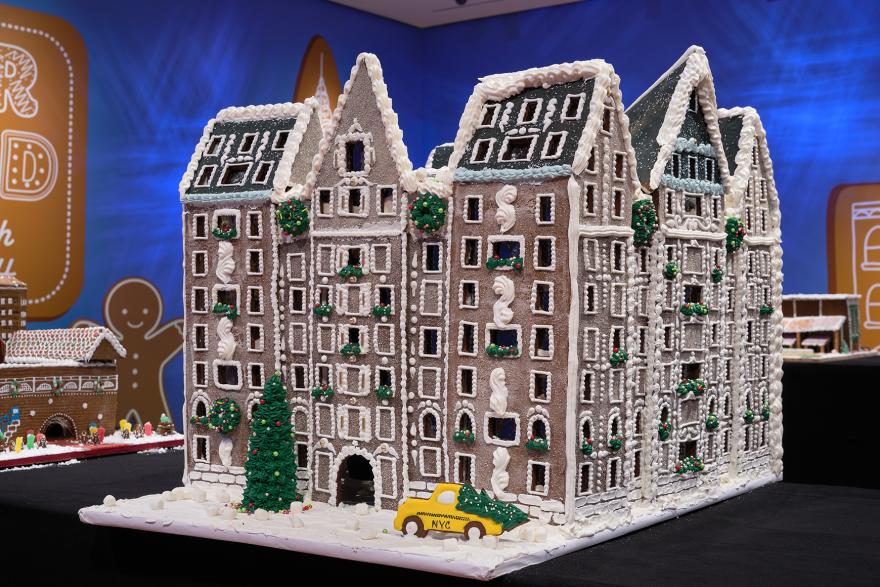 A photograph of a gingerbread house of a large ashy brown hotel decorated with white piping and Christmas-themed symbols, including a Christmas tree larger than the hotel’s entrance, wreaths, and a NYC yellow taxi that has a Christmas tree in its trunk.