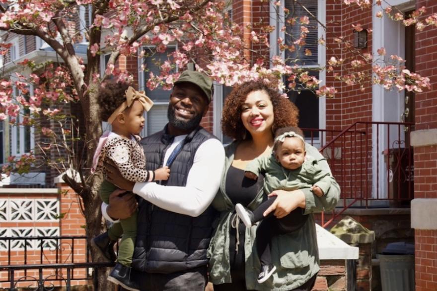 A man and woman, each holding a young child, stand smiling next to each other in front of a blossoming tree and brick building.
