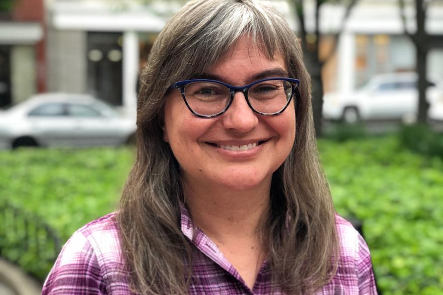 Headshot of a smiling woman wearing dark blue reading glasses and a plaid, purple shirt outdoors.