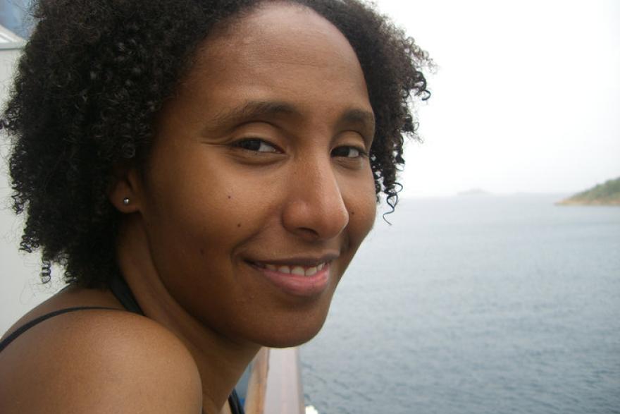 Close-up headshot of a smiling woman on a boat facing the camera with open water and small slivers of land in the background.