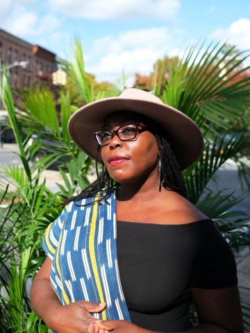 A woman with glasses poses in a hat in front of a large plant.