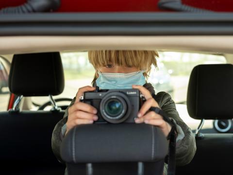A young boy in a mask holds up a camera in the back of a car. 
