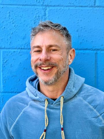 A man smiles at the camera in front of a blue wall. 