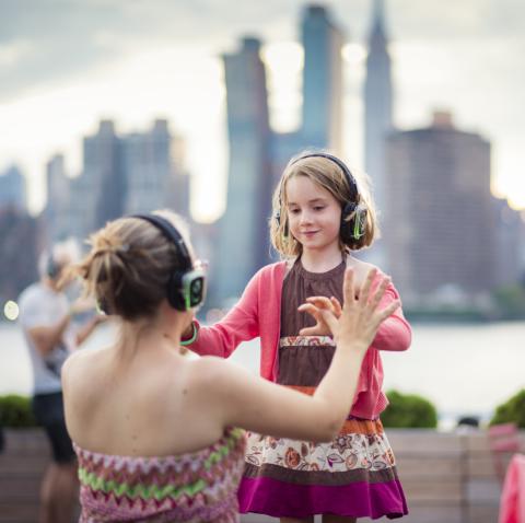 A young girl and her mother, each wearing headphones, are holding hands and dancing outside with the city skyline as a backdrop.