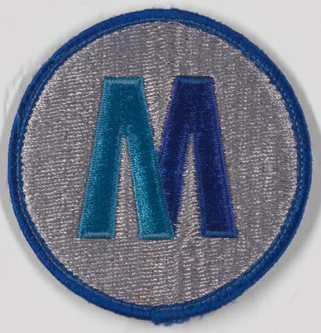 Patch circular de tecido com uma letra maiúscula “M” em dois tons de azul em um campo branco, aparado em azul.