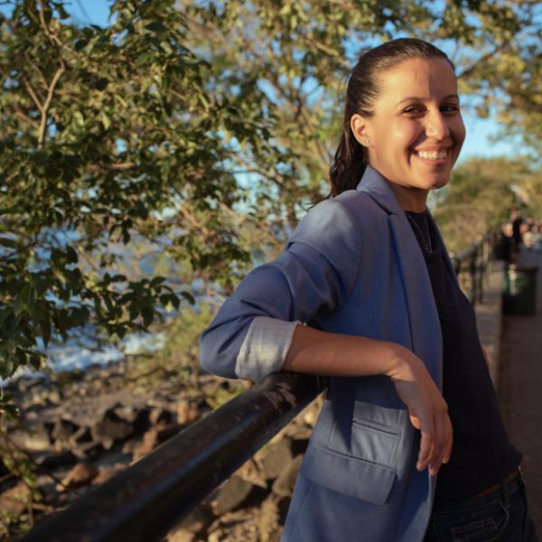 Tiffany Cabán sorri e se inclina em frente a uma ponte do parque. Ela está vestindo um blazer cinza azulado e uma blusa preta. Seu cabelo está preso em um rabo de cavalo.