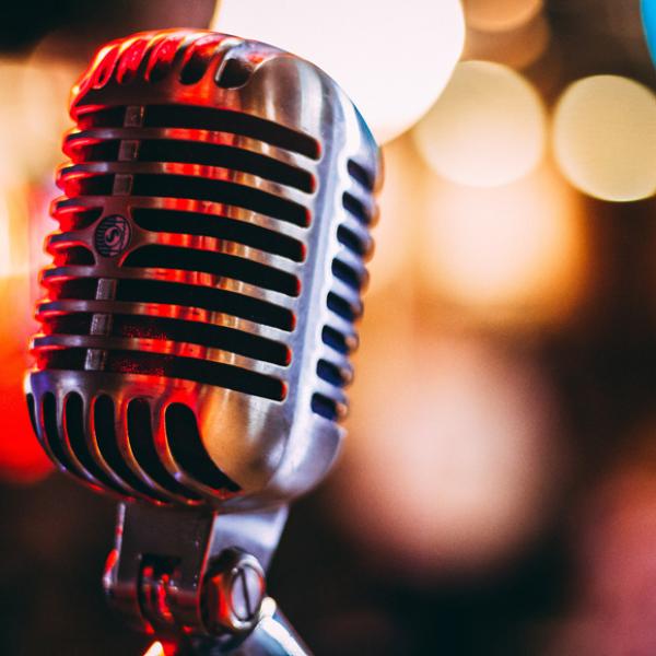 A silver microphone in focus against a blurry background. There are out of focus yellow, red, and blue lights in indistinguishable shapes in the background. 
