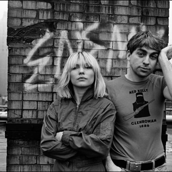 Black and White photograph of a man and woman standing on a roof, facing the camera. The woman has her arms crossed, the man holds a hand up to his temple.