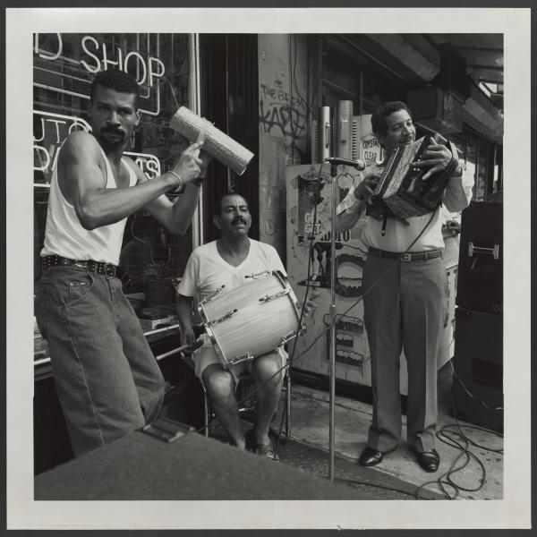 Fotografía en blanco y negro de tres hombres tocando instrumentos musicales fuera de una tienda. Un hombre está sentado con un tambor en el regazo, mientras que los otros dos están de pie: uno rasguea una güira y el otro sonríe mientras toca su instrumento.