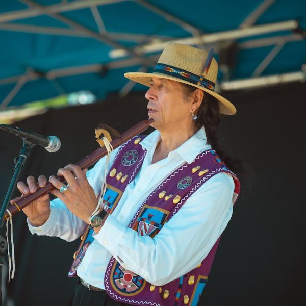 Um homem com um chapéu grande e colete de estilo tradicional toca flauta.