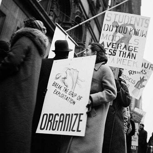 Photograph depicts models for the garment industry protesting working wages out of the Art Students League. The image is black and white and has protestors holding signs that read “Organize,” and “Break the grip of exploitation” 