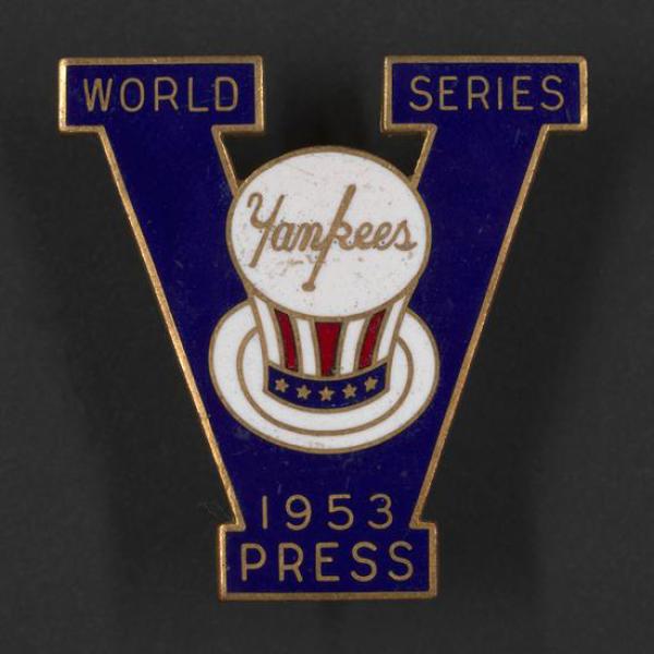 Enamel pin in the shape of the letter “V.” Top left of “V” says “World,” and top right says “Series.” At center is Yankees logo on stars and stripes top hat. At the bottom, “1953, Press.”