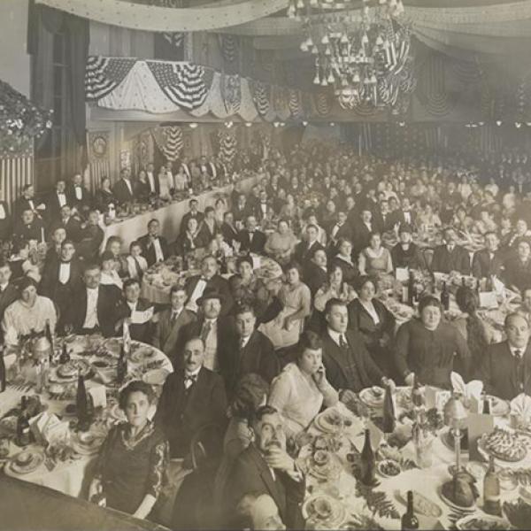 Black and white circa 1910 photograph of a formal dinner banquet. Men, women, and a few children sit at tables looking at camera, place settings, dessert, and wine bottles are visible on the tables.