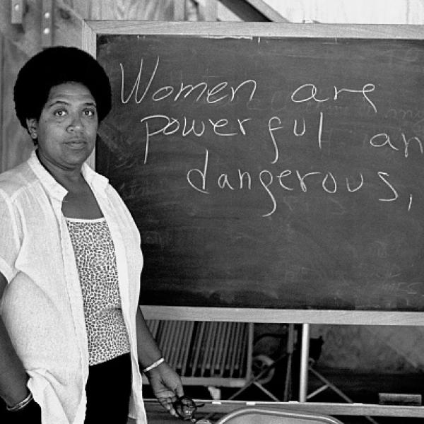 Une photo en noir et blanc d’une femme noire regardant fixement l’appareil photo. L'enseignement d'Audre Lorde, debout à côté d'un tableau noir sur lequel on peut lire "Les femmes sont puissantes et dangereuses".